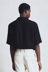 Camisa negra de manga corta en tejido arrugado. Modelo con cuello abierto, tapeta lisa, bolsillo superior y bajo recto. Corte estándar.