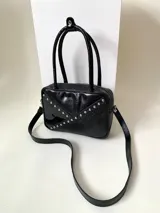 Cartera rígida negra de cuero con tachas plateadas y tres combinaciones de cuero diferentes que aportan textura. Tiene herrajes y cierre metálico plateado marca YKK. Cuenta con un asa fina para llevar al hombro o en la mano y una correa larga extra regulable.