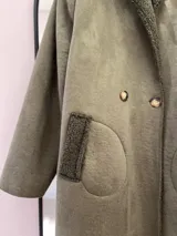 Tapado largo color verde militar, con exterior de gamuza e interior de corderito. Tiene cuello clásico, cierre frontal con doble botonadura y bolsillos de parche redondeados.