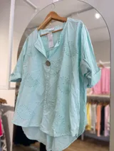 Blusa color crema con cuello camisero, mangas cortas con dobladillo y cierre frontal con un botón de madera. Presenta bordados con motivos de paisley y perforaciones.