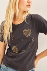 Remera básica de jersey color gris oscuro, con escote redondo y manga corta. Presenta bordados de corazones y estrellas doradas en el frente.