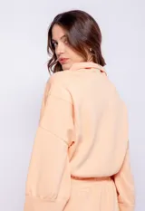 Buzo de tela deportiva color melón, con cuello alto y cierre frontal, manga larga y elásticos en los bajos.
