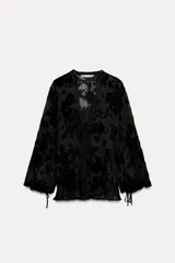 Blusa negra semitransparente de cuello redondo y escote pico ajustable con lazadas. Manga larga abullonada ajustable con cordones. Detalle de aplicación de terciopelo a tono.