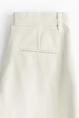 Jean tobillero de denim de algodón rígido color beige claro, con corte estándar de cadera a muslo y piernas con volumen de diseño curvo que se ciñen en el bajo. Presenta tiro alto, cierre con botón, bolsillos al bies, bolsillos decorativos traseros y pliegues delanteros y traseros.