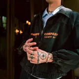 Chaqueta de jean negro con efecto lavado, corte recto, cuello clásico, cierre frontal con cremallera, bolsillos laterales y puños elásticos. Estampado con el texto "VENEFO COMPANY" en color naranja.