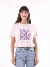 Remera color rosa claro con estampado cuadrado en el frente con motivos florales y manos en tonos lila y naranja.