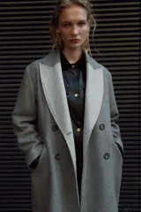 Abrigo gris de corte recto, largo hasta la rodilla, con cuello solapa, doble botonadura frontal y bolsillos con solapa.