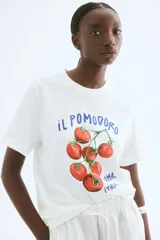 Remera blanca de corte holgado con estampado de tomates y texto en azul. Tiene cuello redondo ribeteado acanalado y hombros caídos.