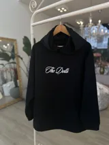 Hoodie negro de corte oversize, con capucha, bolsillo frontal y estampado blanco en la espalda con la inscripción "The Dolls The Dolls VIP CLUB".