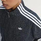 Campera deportiva Adidas Originals modelo Firebird, color negro con estampado animal print sutil. Presenta cuello camisero, cierre frontal y las icónicas tres tiras blancas en las mangas. Lleva el logo del trébol bordado en blanco en el pecho.