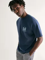 Camiseta azul de manga corta con cuello redondo y estampado frontal con palmeras y texto.