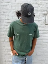 Remera de algodón color verde oscuro con efecto gastado (acid wash). Presenta un estampado minimalista en el pecho con el texto "ON THE MOVE" y un patrón de cuadros blancos a la derecha.