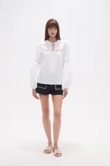 Blusa holgada de algodón color rosa pálido, con escote redondo y lazo ajustable en el centro. Presenta detalles de encaje calado alrededor del escote y en los puños de las mangas largas abullonadas.