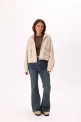 Campera corta color beige claro, estilo bomber o trucker, confeccionada en símil cuero suave. Presenta cuello mao, cierre frontal metálico, bolsillos frontales con tapa y botones a presión, y reguladores ajustables en el ruedo.