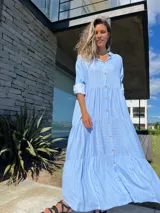 Vestido camisero largo celeste con rayas verticales blancas, cuello camisero, manga larga con puño y abotonadura frontal. Diseño escalonado con volantes en cada corte.