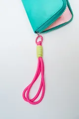 Billetera rectangular color fucsia con cierre perimetral y parche frontal negro con logo. Incluye correa de mano de cordón rosa neón y detalle de ajuste en verde lima.