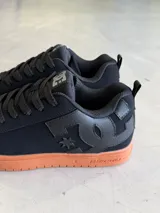 Championes DC Court Graffik color negro con suela de goma marrón. Presentan cordones gruesos negros, logo de DC en la lengüeta y en el lateral, y detalles de costuras.