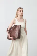 Cartera tipo tote bag en símil cuero color marrón oscuro, con bordados decorativos en blanco que evocan un estilo western. Cuenta con asas largas para llevar al hombro.