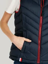 Chaleco acolchado azul marino con capucha ajustable con cordones y cierre de cremallera frontal. Bolsillos laterales con cremallera y logo de Tommy Hilfiger bordado en el pecho. Detalles distintivos de Tommy Hilfiger en el ruedo.