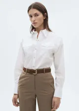 Camisa blanca de algodón con cuello camisero, cierre frontal con botones efecto joya, dos bolsillos de solapa en el pecho, manga larga abotonada y corte recto.
