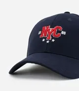 Gorra de sarga de algodón color marrón oscuro con visera curva y ajuste trasero. Presenta un bordado frontal con las letras 'NYC' en estilo universitario con detalles en rojo y blanco, flanqueadas por los números '19' y '86' y tres estrellas blancas debajo.