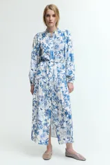 Vestido largo blanco con estampado floral en tonos azules. Tiene cuello mao, botones ocultos en la parte delantera, mangas largas abullonadas con puños elásticos, cinturón fino extraíble y costura fruncida debajo de la cadera para dar vuelo.