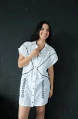 Vestido corto blanco de algodón con cuello camisero, manga corta y cierre frontal con botones. Presenta bordados calados con detalles en negro y ribetes negros en cuello, mangas y cierre.