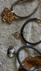 Collar con cordón trenzado de cuero sintético color negro, con dije circular plateado con centro de piedra blanca con pintitas negras. Cierre con mosquetón plateado.