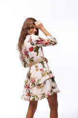 Vestido corto blanco con estampado floral en tonos rojos, verdes y amarillos. Tiene escote en V, mangas largas abullonadas y falda con volados. Se ajusta a la cintura con un cinturón de soga con hebilla circular.