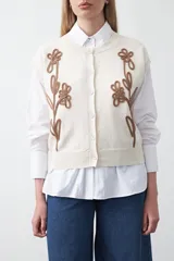 Chaleco de punto color blanco con bordado aplicado de cordón marrón en diseño floral. Presenta cierre frontal de botones al tono, cuello redondo y terminaciones acanaladas en sisas y ruedo.