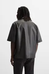 Sobrecamisa negra de manga corta confeccionada en tejido efecto piel, con cuello solapa, bolsillo frontal de parche y cierre frontal con botonadura oculta.