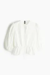 Blusa blanca de algodón con textura de lunares. Tiene escote redondo con lazo, botones en el frente, mangas tres cuartos abullonadas, cordón ajustable en la cintura y peplum.