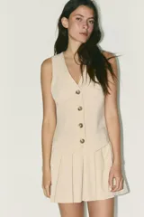 Vestido corto color beige, de escote pico y sin mangas. Tiene falsos bolsillos de vivo en el delantero, falda con tablas y detalle de trabilla en la espalda. Cierre lateral con cremallera oculta.