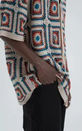 Camisa de manga corta, tejida a crochet con un patrón de cuadrados en colores beige, naranja y turquesa. Presenta cuello tipo polo y cierre frontal con botones.