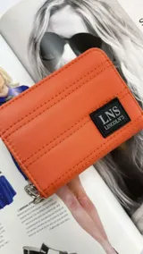 Billetera corta con cierre alrededor y tapa, confeccionada en tela capitoneada. Incluye varias reparticiones con tarjeteros y bolsillos internos, y una correa de mano desmontable. Disponible en negro, rojo y dorado.