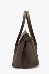 Bolso marrón de gamuza sintética, tipo city bag, con doble asa de hombro y cierre con solapa e imán. Tiene correa ajustable con hebillas metálicas en los laterales y bolsillo interior con cremallera.