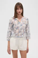 Blusa blanca de algodón con bordado broderie inglés y estampado floral en tonos azules y marrones. Posee cuello en V con abertura y botones frontales, y mangas tres cuartos.