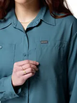 Camisa de senderismo de manga larga color azul verdoso, con protección solar FPS 50, tejido absorbente de sudor y ventilaciones de malla. Cuenta con dos bolsillos frontales con cierre, tapeta abotonada y lazo para colgar.