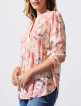 Blusa de gasa estampada con flores en tonos rosa, amarillo y azul. Tiene cuello a la base con escote en V y botones. La parte delantera tiene detalle plisado y las mangas son 3/4 regulables con presilla y botón.