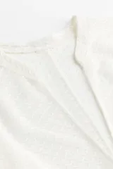 Túnica corta blanca de punto liviano con cuello corto y abertura en V adelante. Modelo de corte holgado con hombros caídos, mangas largas holgadas y bajo recto.