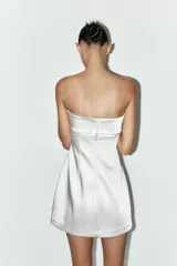 Vestido corto blanco de satén, con escote recto palabra de honor, detalle de lazo en la espalda y cierre trasero con cremallera oculta.