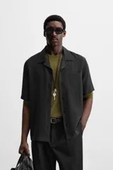 Camisa negra de corte relaxed fit, confeccionada en tejido con mezcla de modal. Tiene cuello bowling, manga corta, bolsillo de parche en el pecho y cierre frontal con botones.