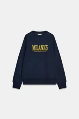 Sudadera de tejido de punto color azul marino, con cuello redondo y mangas raglán. Presenta un estampado frontal en amarillo con el texto "MILANO 75" y una dirección debajo.