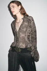 Camisa de viscosa con estampado paisley en blanco y negro. Cuello redondo con escote pico ajustable con cordones, mangas largas con elástico y volantes en los puños, bajo asimétrico y cierre frontal con botones forrados.