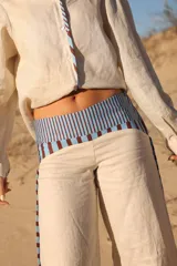 Pantalón de lino color crudo, de corte ancho y tiro medio. Presenta detalles de rayas verticales en color marrón y celeste en los laterales y en la cintura.