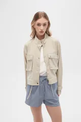 Campera color beige de manga larga, con cuello alto, cierre frontal con cremallera y botones a presión, bolsillos con solapa en el pecho y puños con trabilla ajustable.
