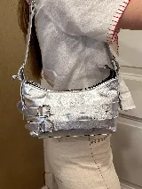 Cartera bandolera marrón de cuero con correa de hombro ajustable, cierre superior con cremallera y detalles de hebillas y aros.