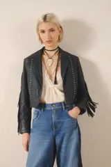 Campera corta de cuero color negro, con flecos en espalda y mangas, tachas metálicas y símil piedra cristal. Cierra con broches en el frente, tiene forrería y bolsillo interno.
