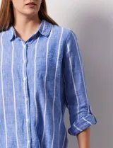 Camisa color beige con rayas verticales blancas, de lino, con cuello con solapa y abertura central con botones. Tiene un bolsillo aplicado en el pecho y mangas largas regulables con presilla y botón.