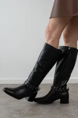 Bota de cuero color marrón con caña alta, taco cuadrado y punta fina. Presenta una correa con ojales metálicos dorados alrededor del tobillo.